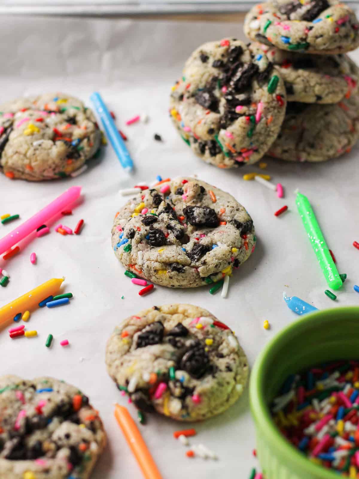 Cookies with rainbow sprinkles and Oreo pieces, sitting on a white piece of parchment and surrounded with birthday candles pointing towards a few of them.