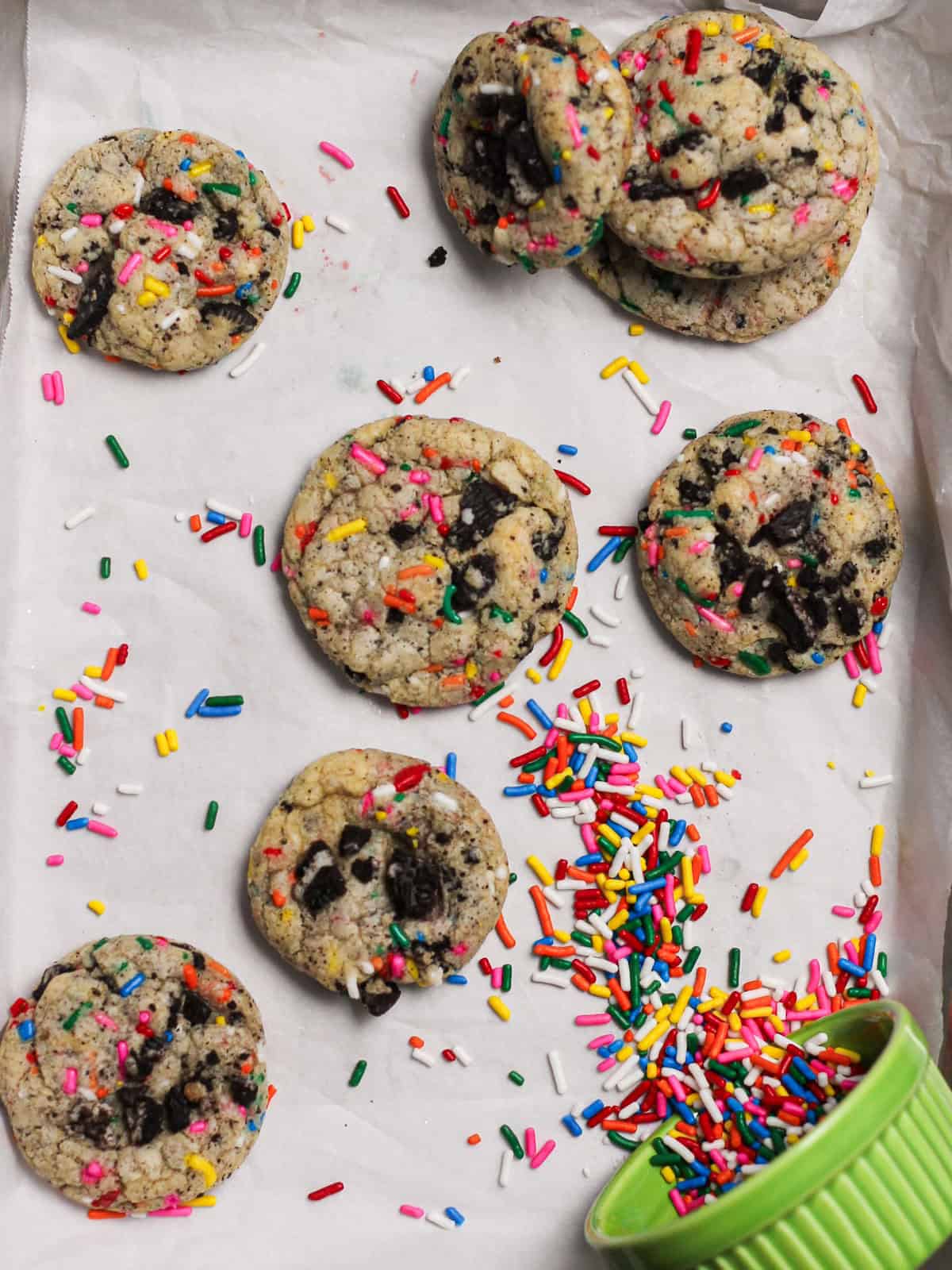 Cookies with rainbow sprinkles and Oreo pieces, sitting on a white piece of parchment.