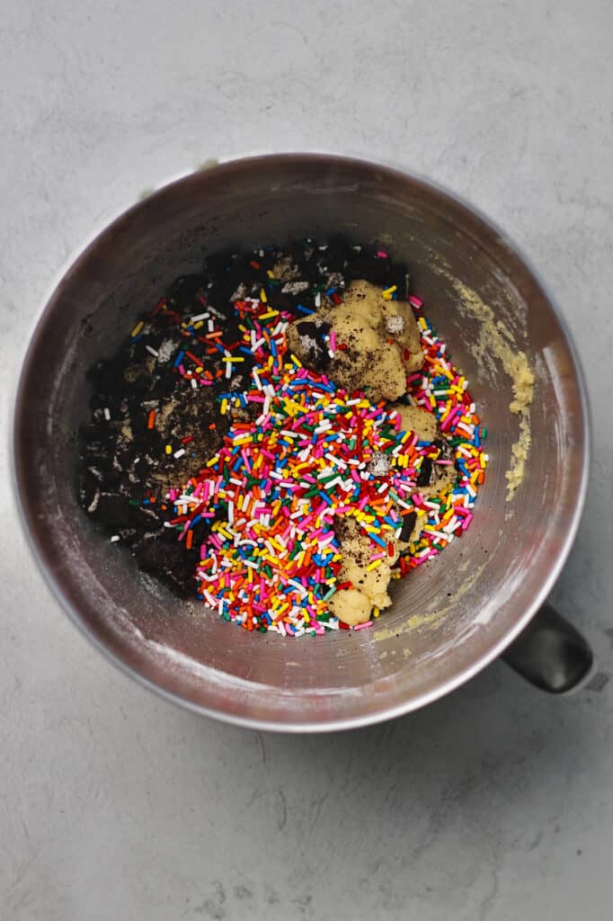 mixing bowl full of light colored cookie dough, filled with rainbow sprinkles and crushed Oreo pieces