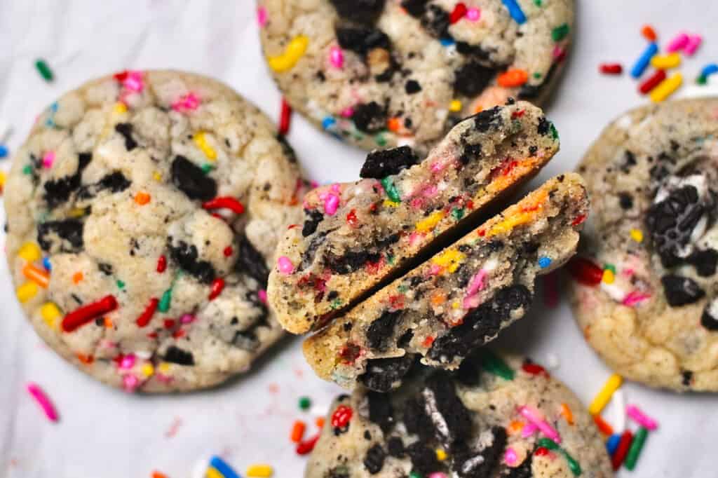 Oreo Funfetti cookie broken in half so that the inside is facing up toward the camera and is surrounded by other similar cookies and sprinkles