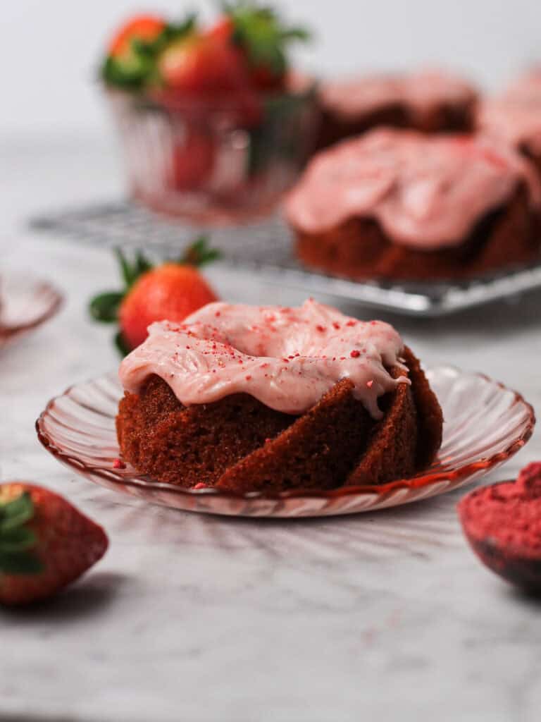 Small strawberry Bundt cake sitting on a little pink plate, surrounded by strawberries and coated with strawberry cream. cheese icing.