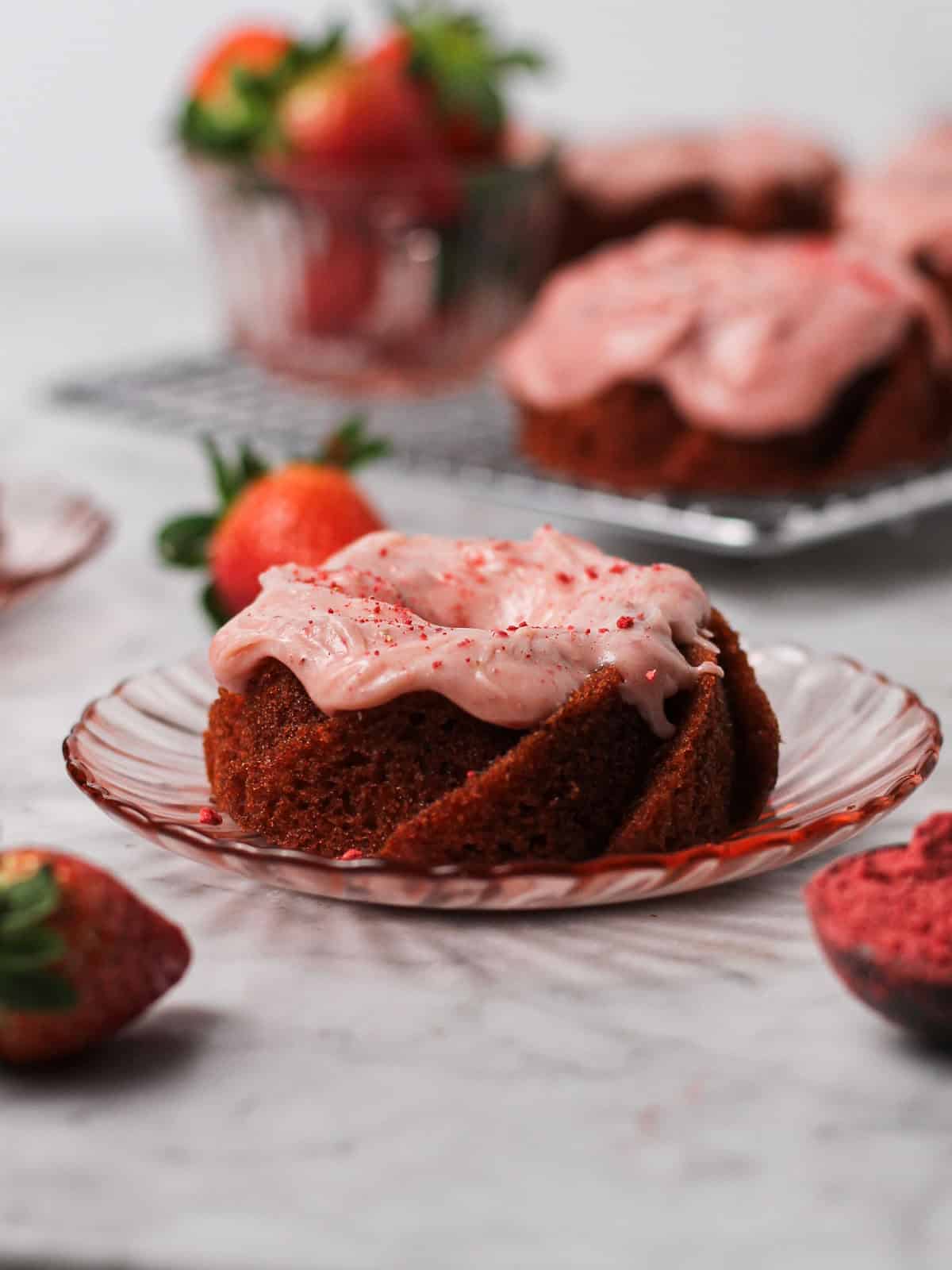 Small strawberry Bundt cake sitting on a little pink plate, surrounded by strawberries and coated with strawberry cream. cheese icing.