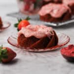 Small strawberry Bundt cake sitting on a little pink plate, surrounded by strawberries and coated with strawberry cream. cheese icing.