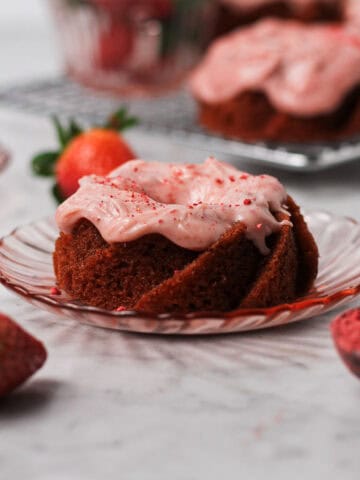 Small strawberry Bundt cake sitting on a little pink plate, surrounded by strawberries and coated with strawberry cream. cheese icing.