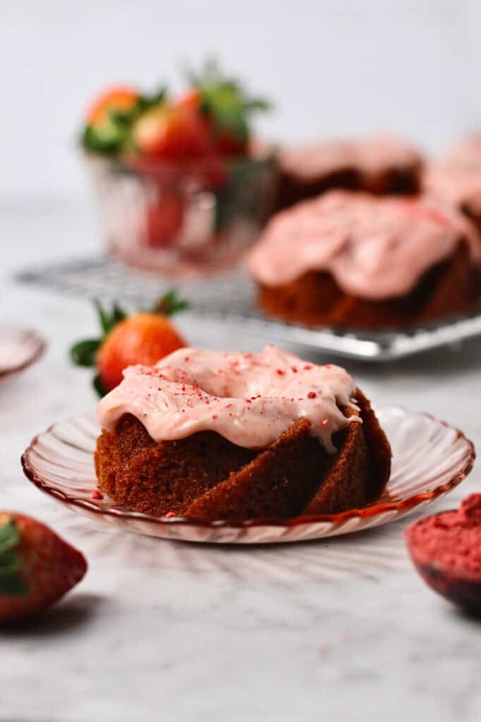 small strawberry cake sitting on a little pink plate, surrounded by strawberries and coated with strawberry cream cheese icing.