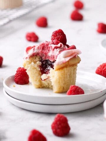 almond raspberry cupcake with a bite missing from it, sitting on stack of small white plates.