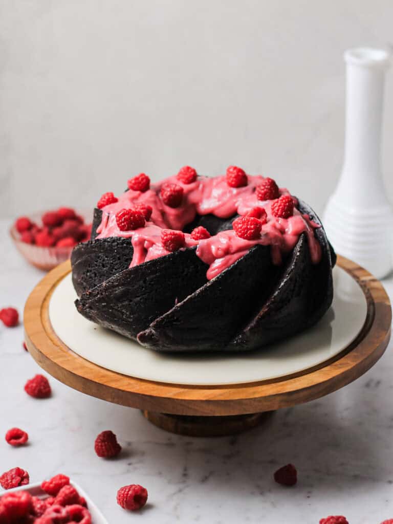 chocolate Bundt cake, sitting on brown wooden cake stand. cake is coated with a pink raspberry cream cheese glaze and fresh raspberries on top.
