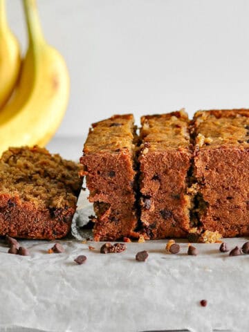 A loaf of chocolate chip oatmeal banana bread from its side as it sittings on a pan, surrounded by mini chocolate chips and bananas in the background.