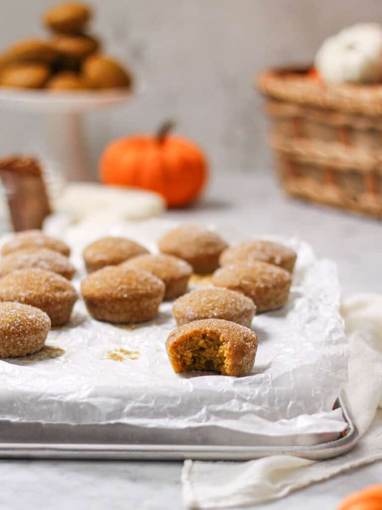 Mini pumpkin muffins sitting on a white piece of parchment paper, with pumpkins and a basket in the background.
