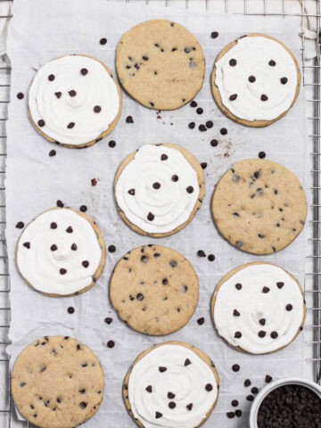 Overhead image of frosted and unfrosted chocolate chip sugar cookies sitting on a wire rack. Parchment paper is on top of the rack and beneath the cookies.