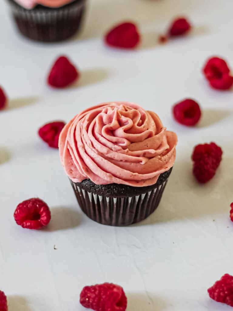 Pink frosting on a chocolate cupcake, surrounded by fresh raspberries.