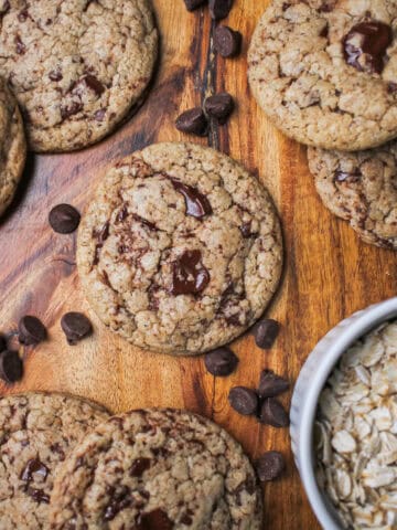 Oat flour chocolate chip cookies, sitting against a brown wooden cutting board and surrounded by chocolate chips.