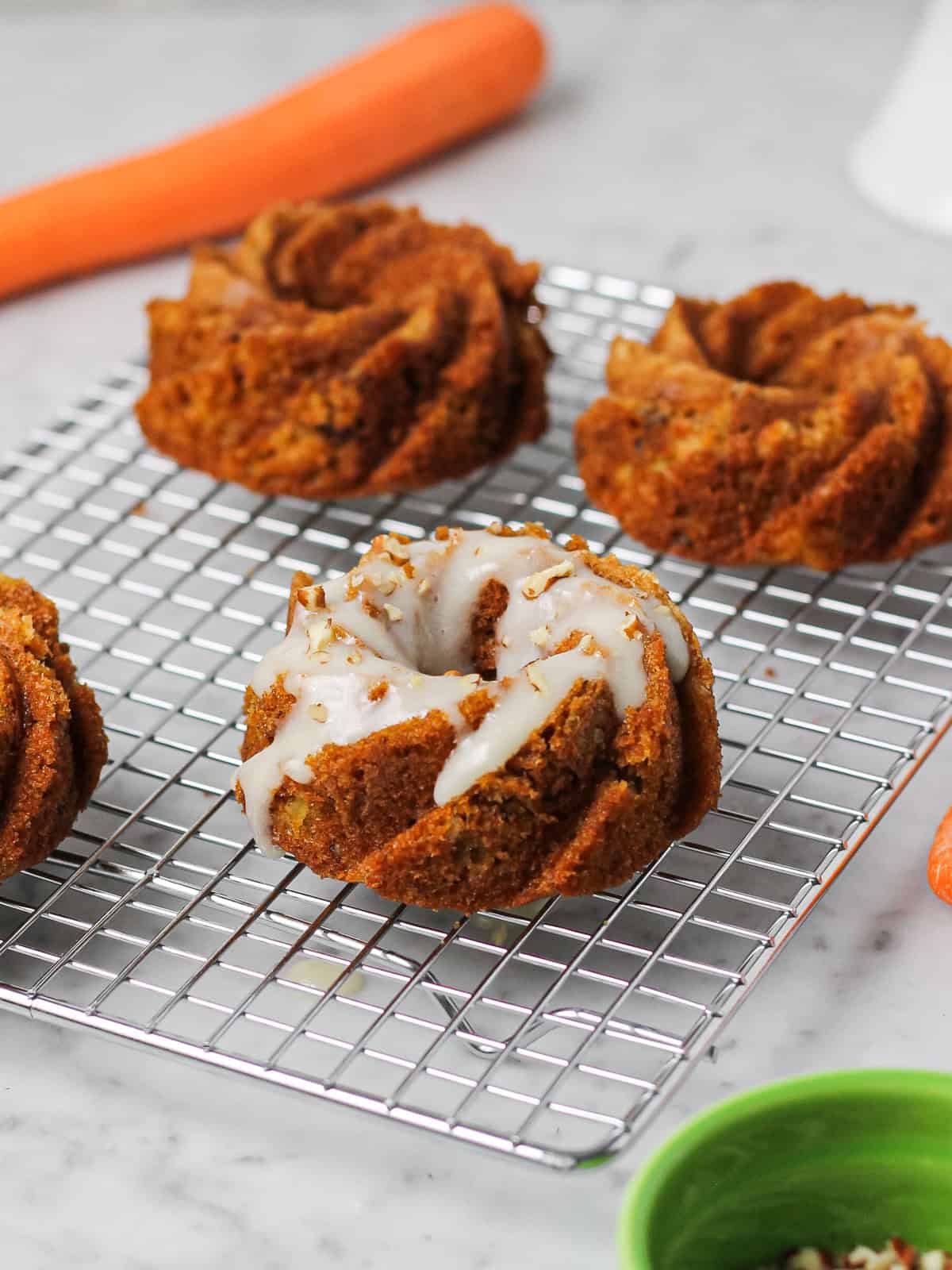 Mini carrot bundt cakes on a cooling rack. Cake is topped with a white cream cheese glaze and nuts, while unglazed mini Bundts sit in the background.