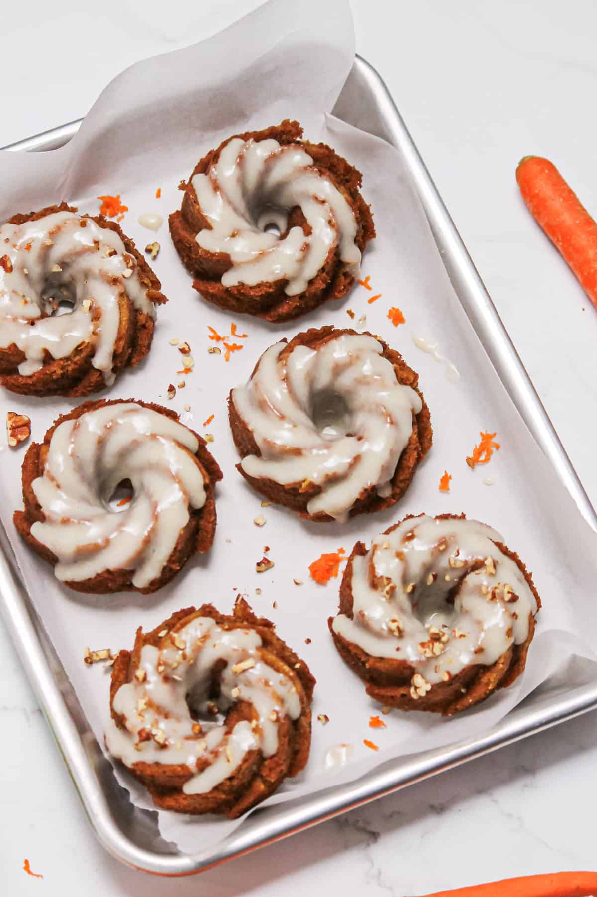 Six mini carrot bundt cakes on a white piece of parchment paper. Cake is topped with a white cream cheese glaze and nuts.
