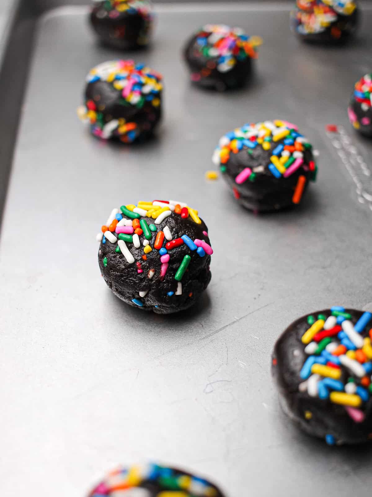 Dough balls of chocolate sprinkle cookies, topped with lots of rainbow sprinkles!