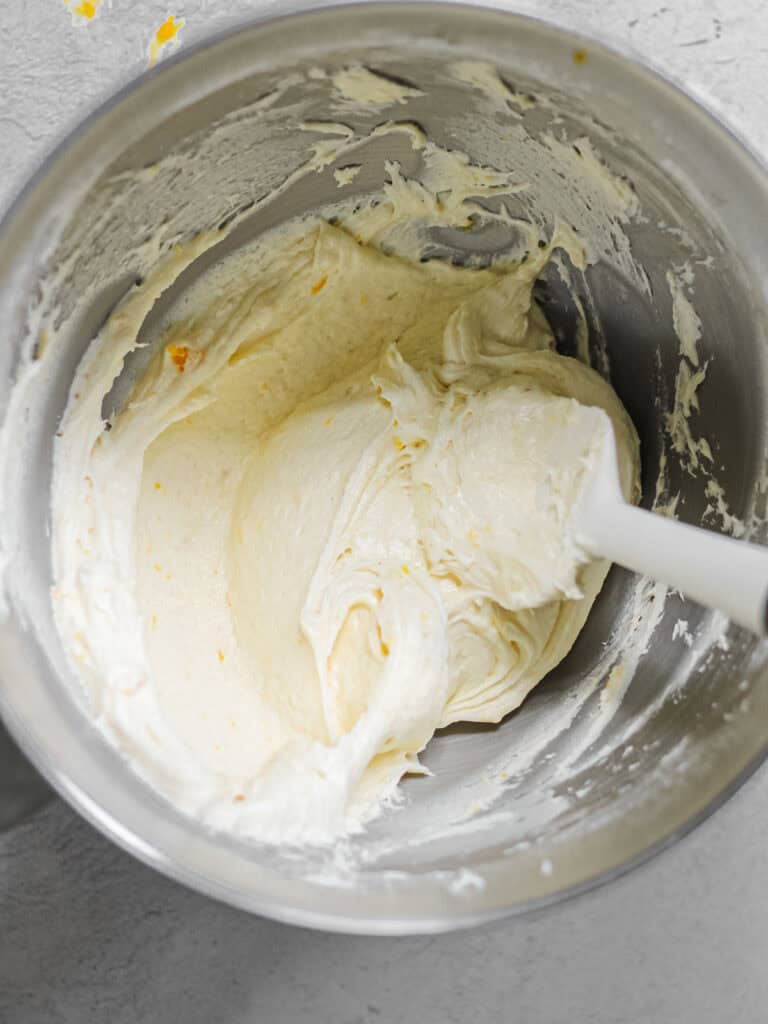 Mixing bowl filled smooth mango buttercream and a white rubber spatula.