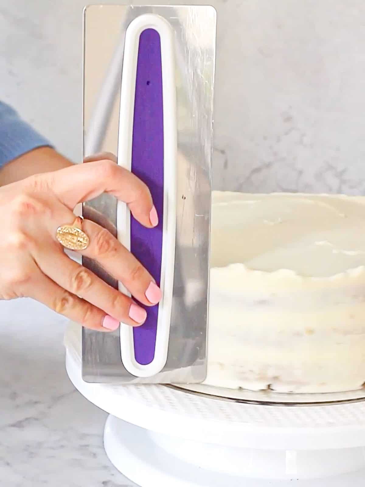 Placing a cake scraper against the side of a layer cake to smooth out its cream cheese frosting.