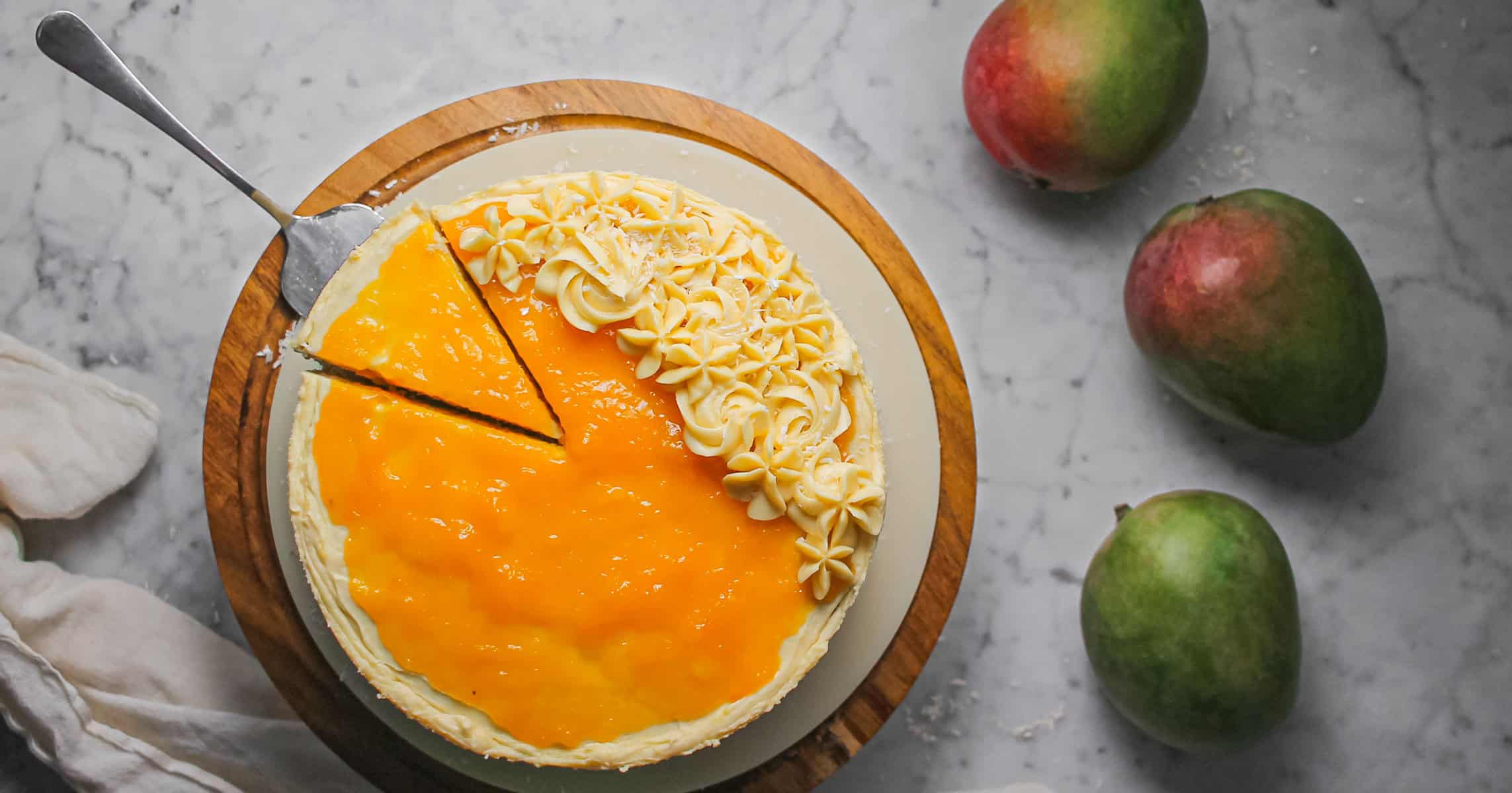 Overhead image mango coconut cake, sitting on a wooden cake stand with three mangoes off to the side.