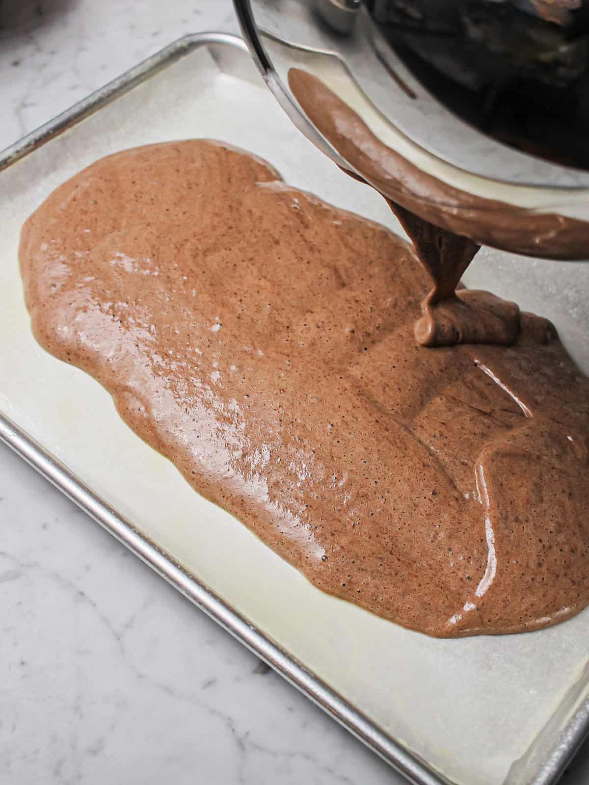 Pouring chocolate sponge batter into a parchment-lined baking sheet.