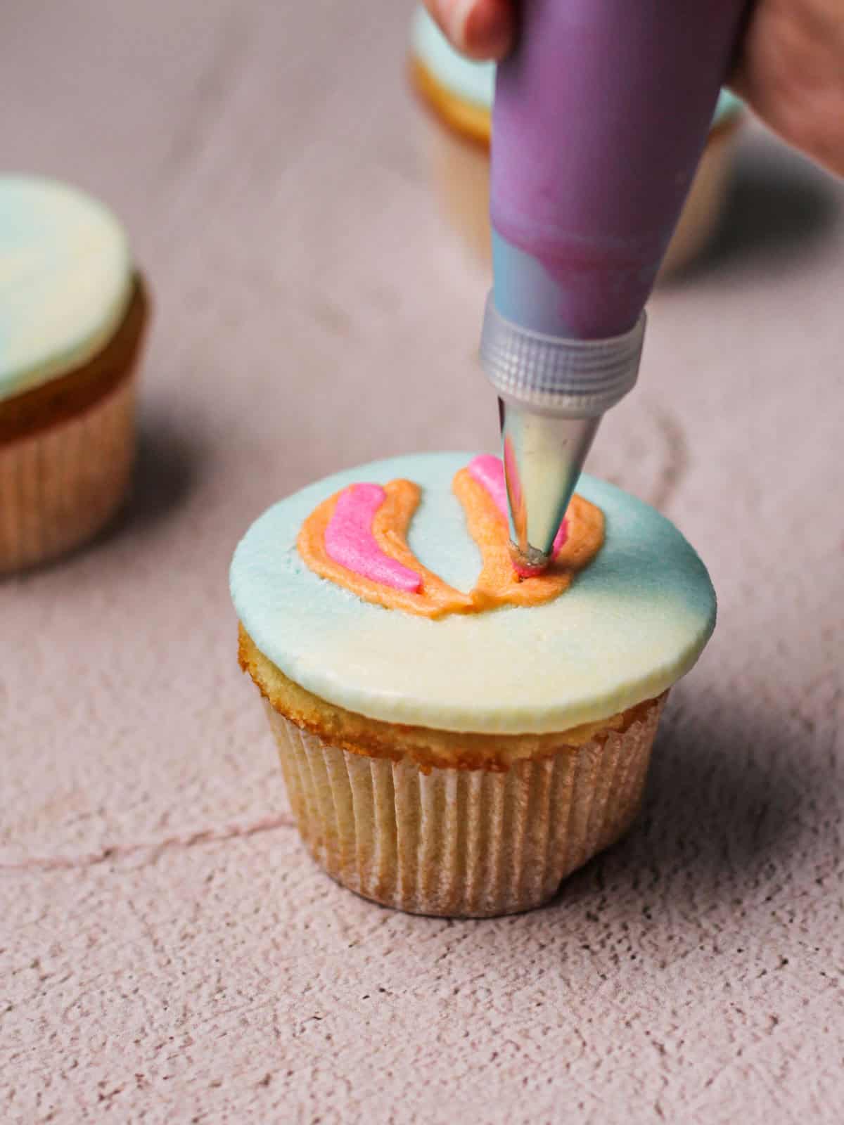 Piping an orange and pink hot air balloon onto a cupcake with frosting.