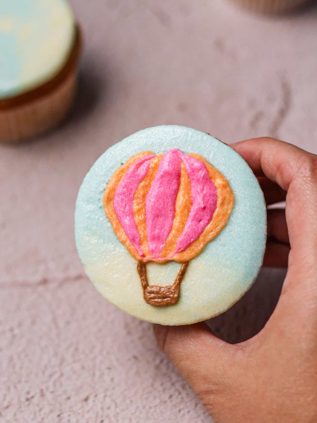 A white hand holding a cupcake with a hot air balloon design.