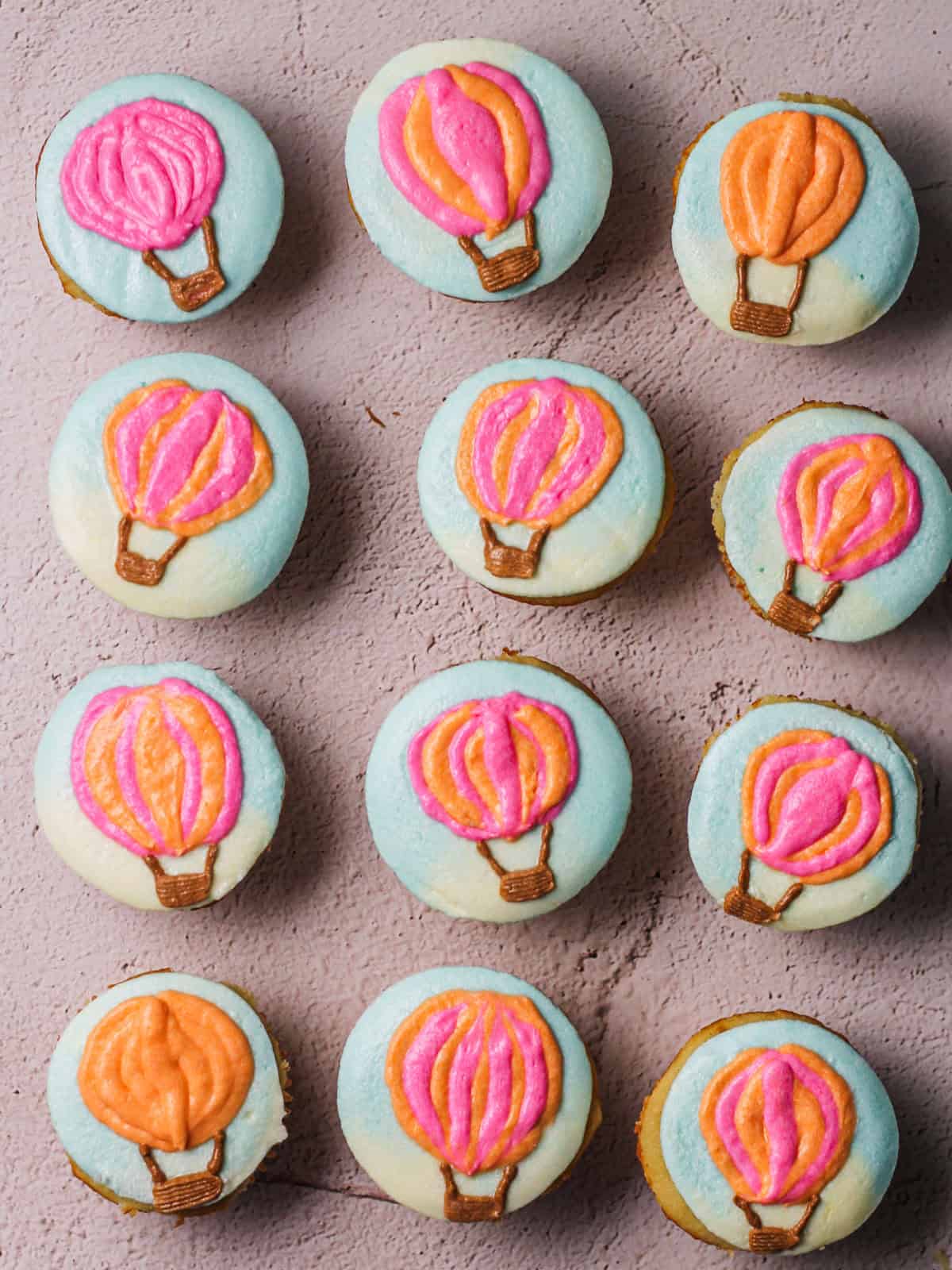 Overhead shot of a dozen hot air balloon cupcakes, with blue frosting as the background and pink and orange balloons.