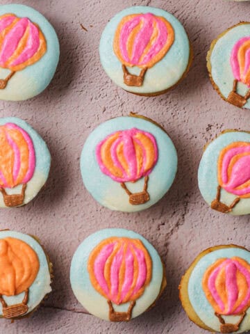 Overhead shot of nine hot air balloon cupcakes, with blue frosting as the background and pink and orange balloons.
