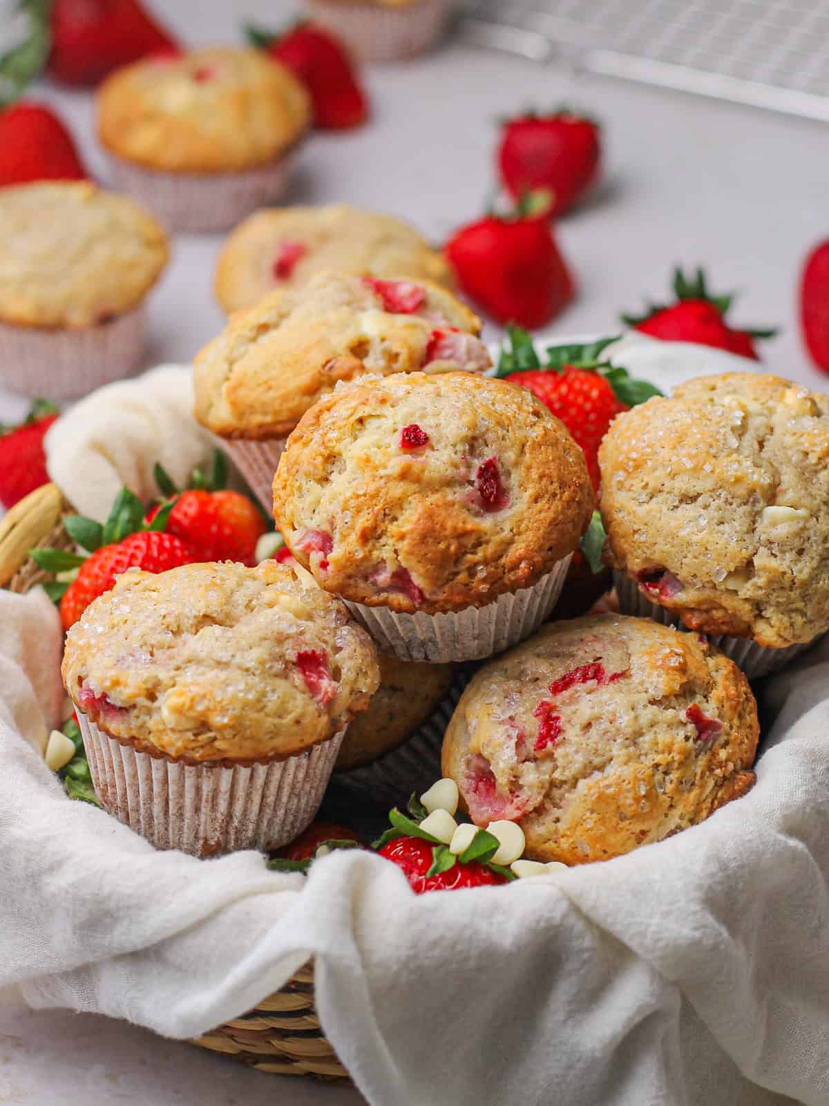White chocolate and strawberry muffins sitting in a basket with strawberries and white chocolate chips peeking out around them.