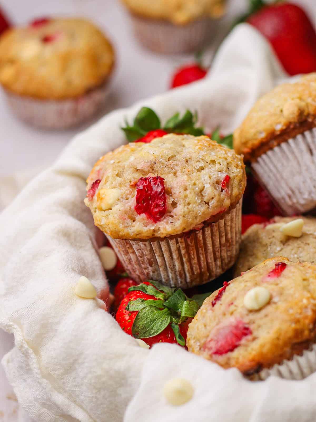 White chocolate and strawberry muffins sitting in a basket with strawberries and white chocolate chips peeking out around them.