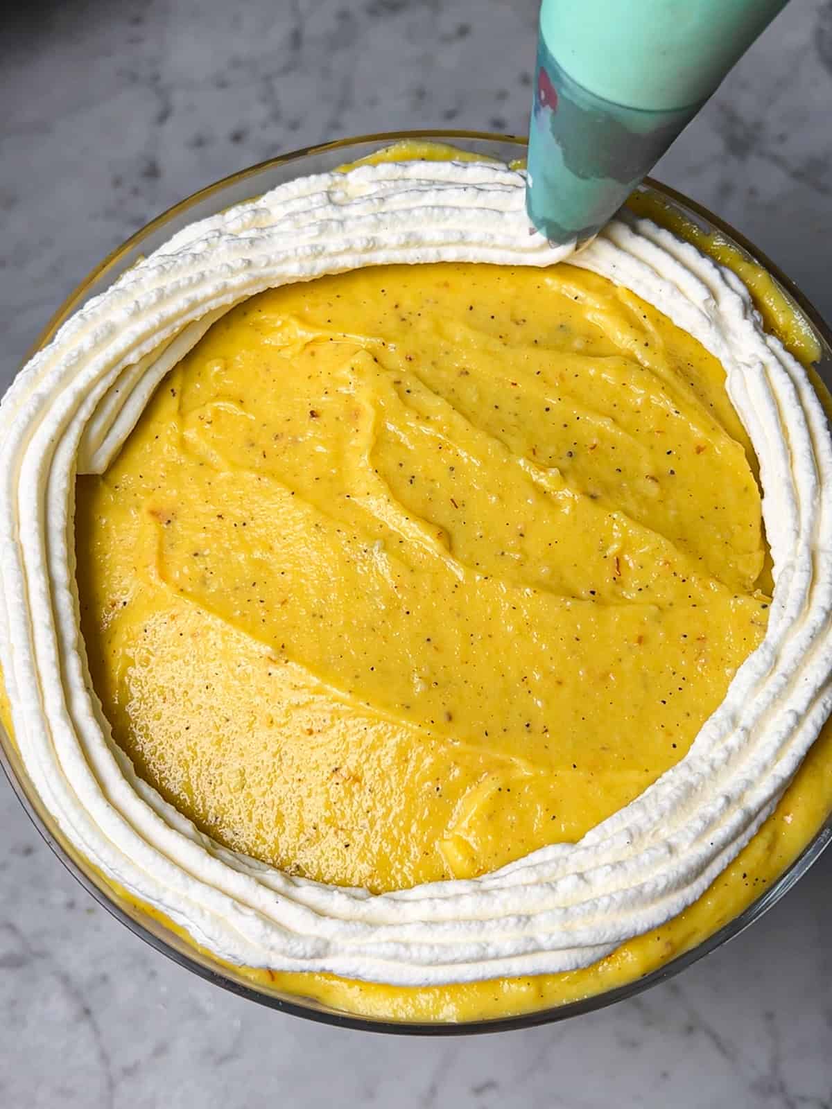 Piping whipped cream onto Saffron phirni in trifle dish.