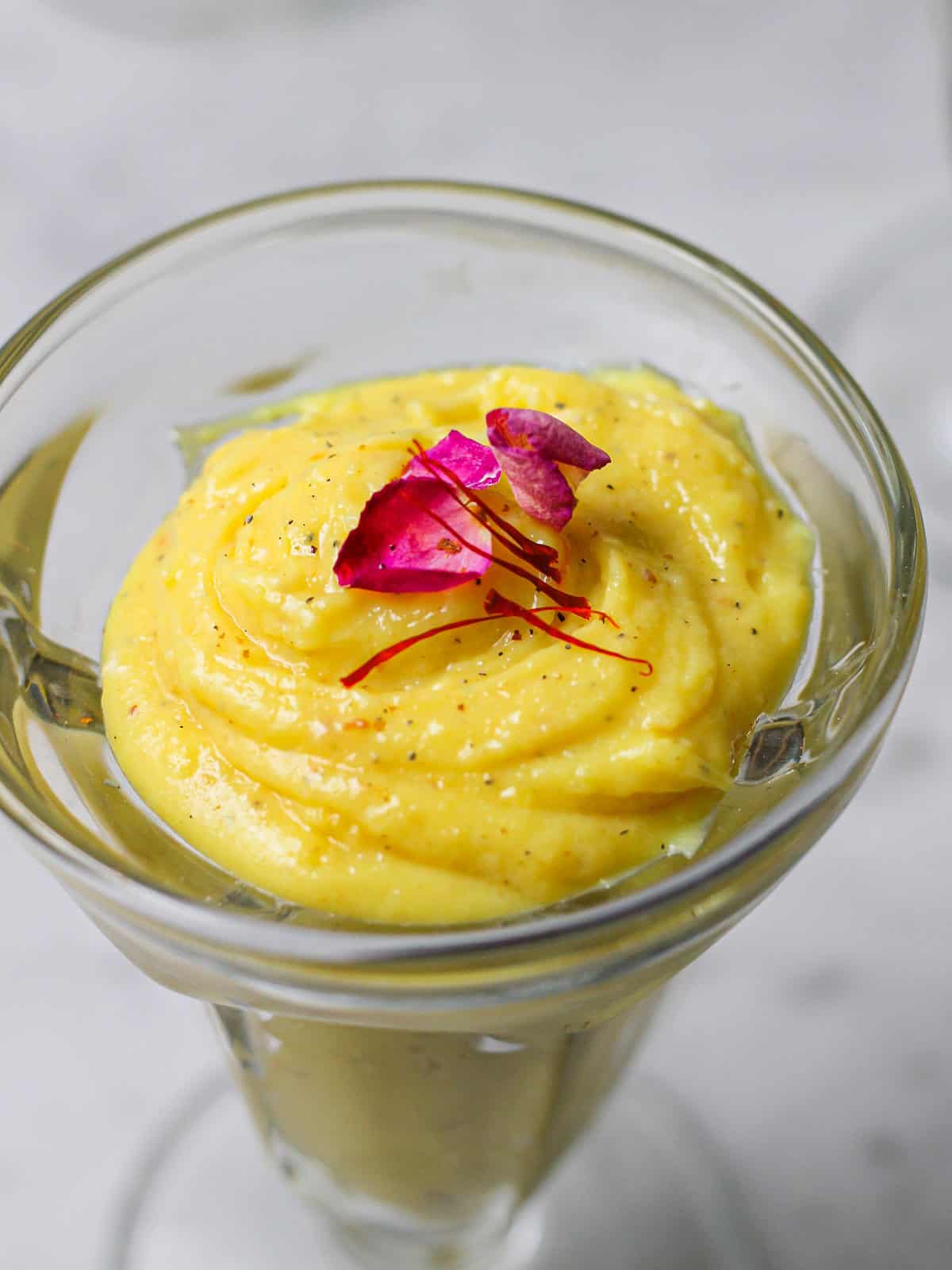 Up-close overhead shot of saffron phirni in a tall glass, with a rose petal and saffron thread on top of the custard.