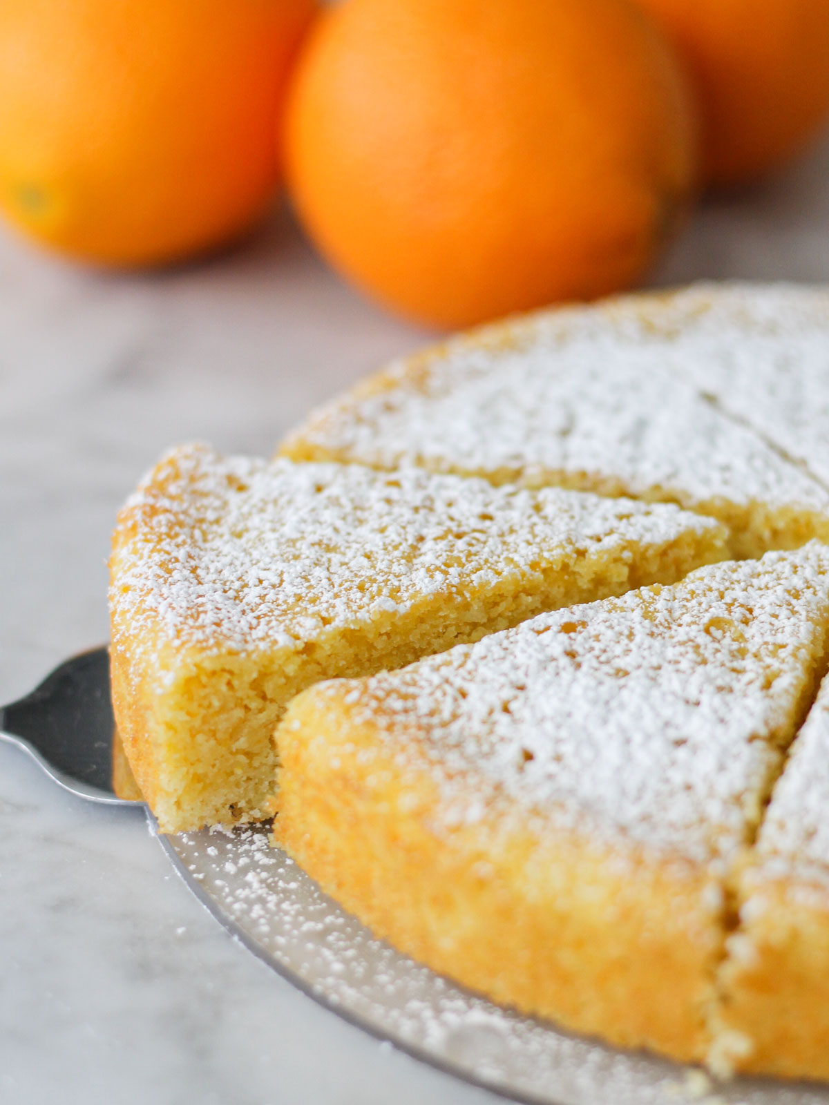 Cake server offset on a cake dish with a slice of Sidi Joon's orange cake on it. Slices are dusted in powdered sugar, with some oranges in the background.