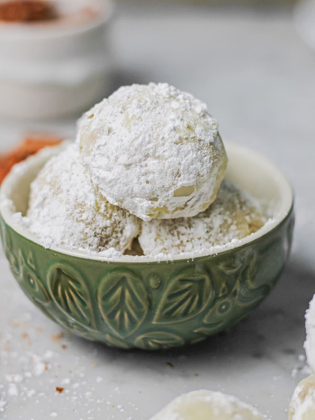 Snowball cookies, dusted in powdered sugar, sitting in a muted green serving bowl, surrounded by bits of pecans.