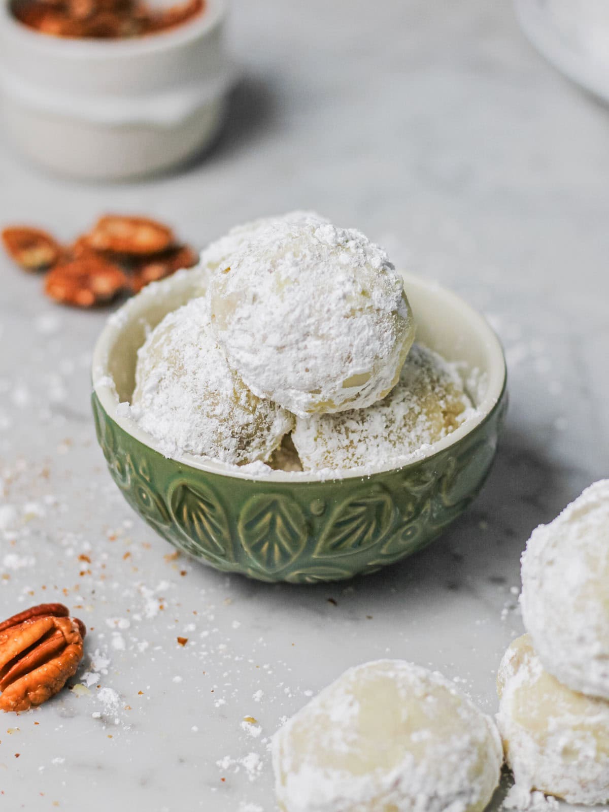 Snowball cookies, dusted in powdered sugar, sitting in a muted green serving bowl, surrounded by bits of pecans.