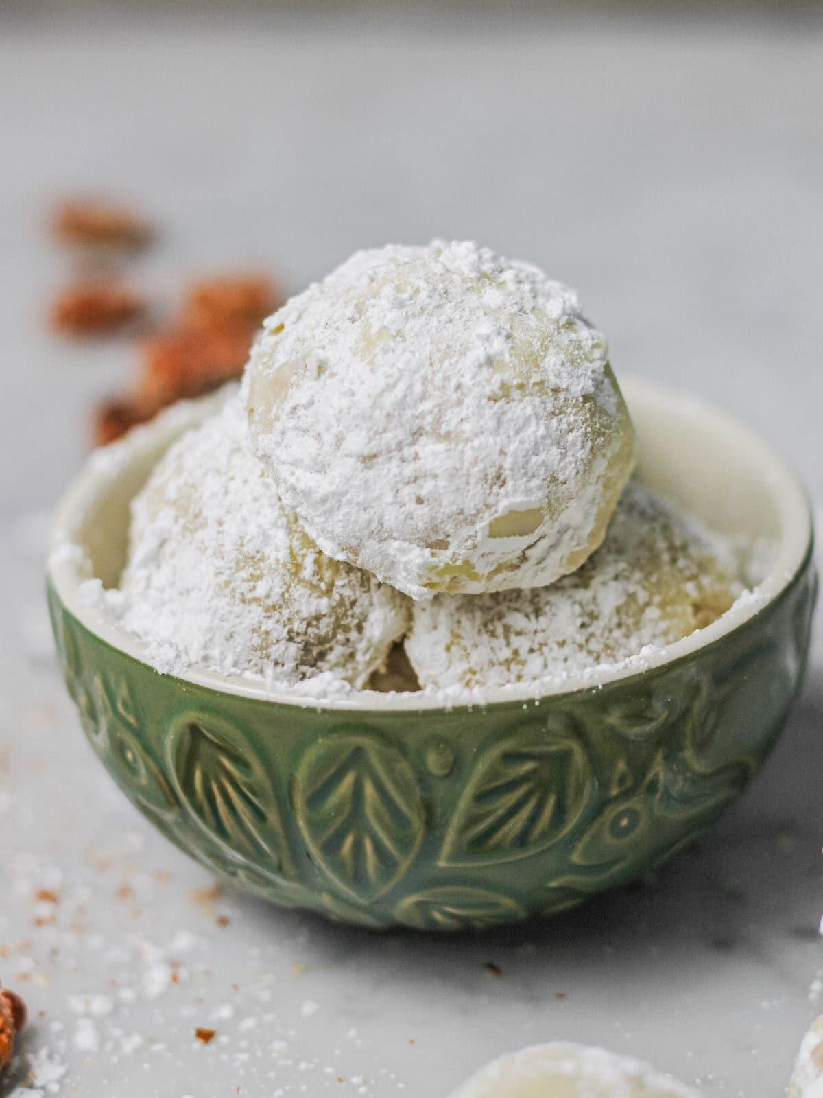 Snowball cookies, dusted in powdered sugar, sitting in a muted green serving bowl, surrounded by bits of pecans.