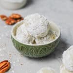 Snowball cookies, dusted in powdered sugar, sitting in a muted green serving bowl, surrounded by bits of pecans.