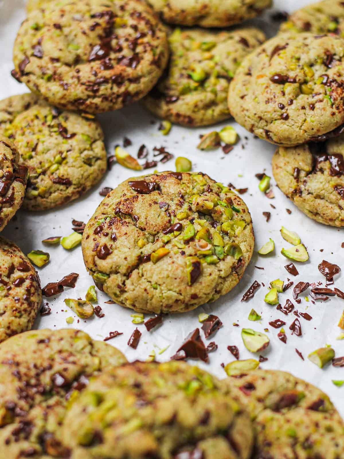 Brown butter pistachio dark chocolate chunk cookies on a white piece of parchment paper, surrounded by chopped pistachios and chocolate chunks.
