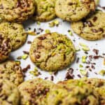 Brown butter pistachio dark chocolate chunk cookies on a white piece of parchment paper, surrounded by chopped pistachios and chocolate chunks.