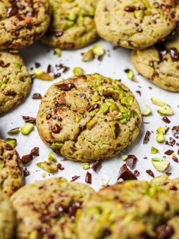 Brown butter pistachio dark chocolate chunk cookies on a white piece of parchment paper, surrounded by chopped pistachios and chocolate chunks.