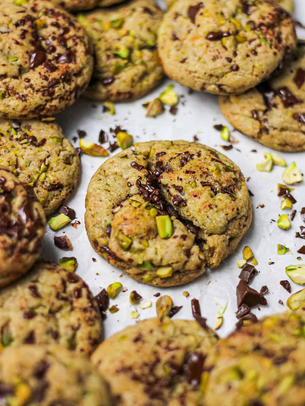 Brown butter pistachio dark chocolate chunk cookies on a white piece of parchment paper, surrounded by chopped pistachios and chocolate chunks.