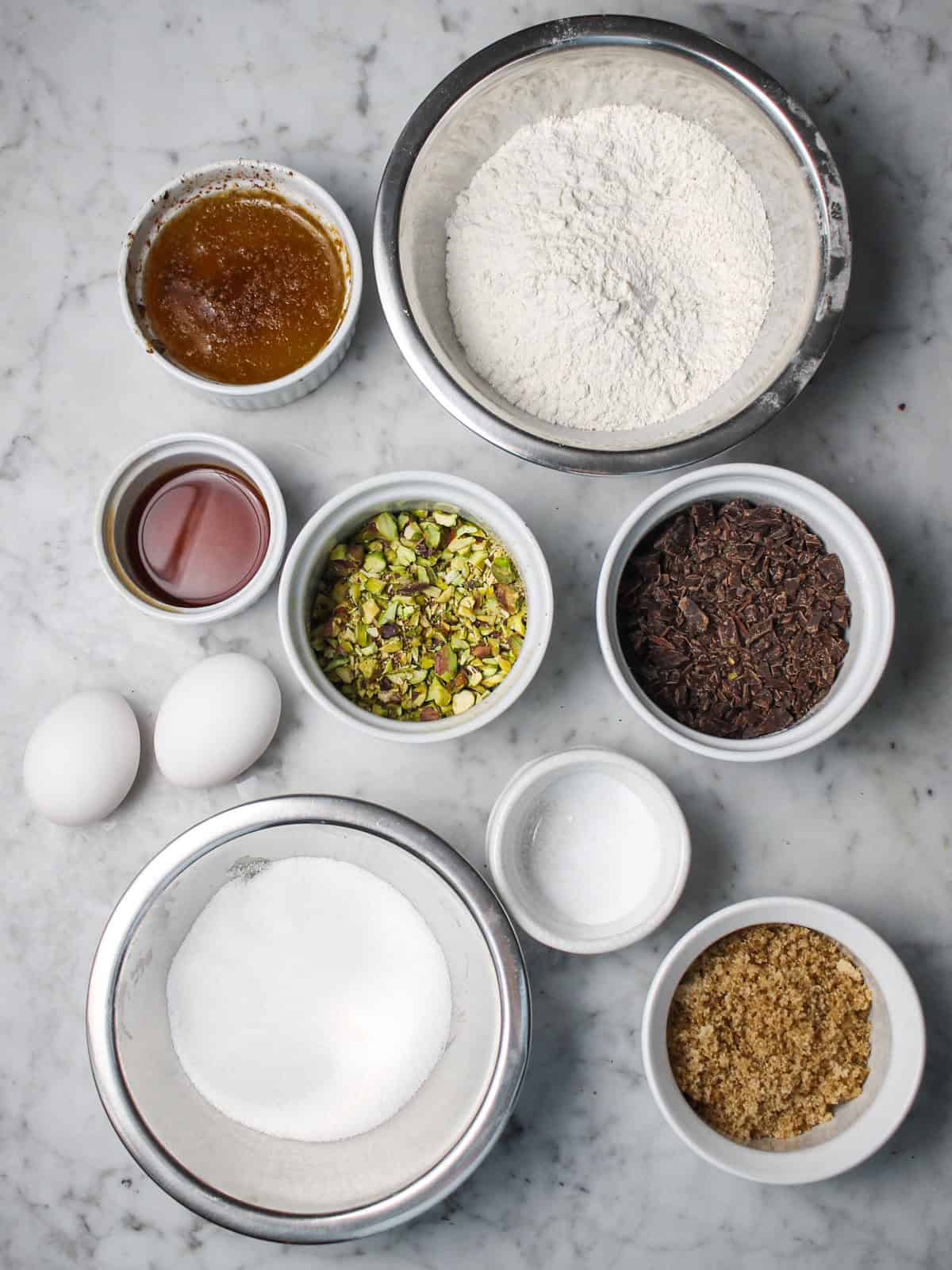 Ingredients including browned butter, pistachios, chopped dark chocolate, eggs, and flour on a countertop.