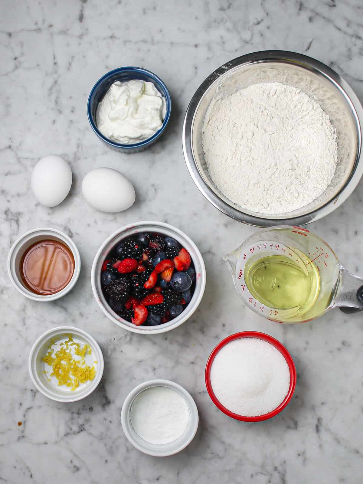 Ingredients for mini mixed berry loaf cakes, including fresh berries, flour, Greek yogurt, eggs, and sugar.