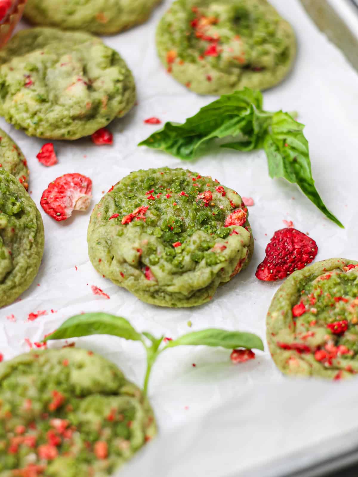 45 degree angle photo of strawberry basil sugar cookies, surrounded by basil leaves and freeze-dried strawberry pieces.