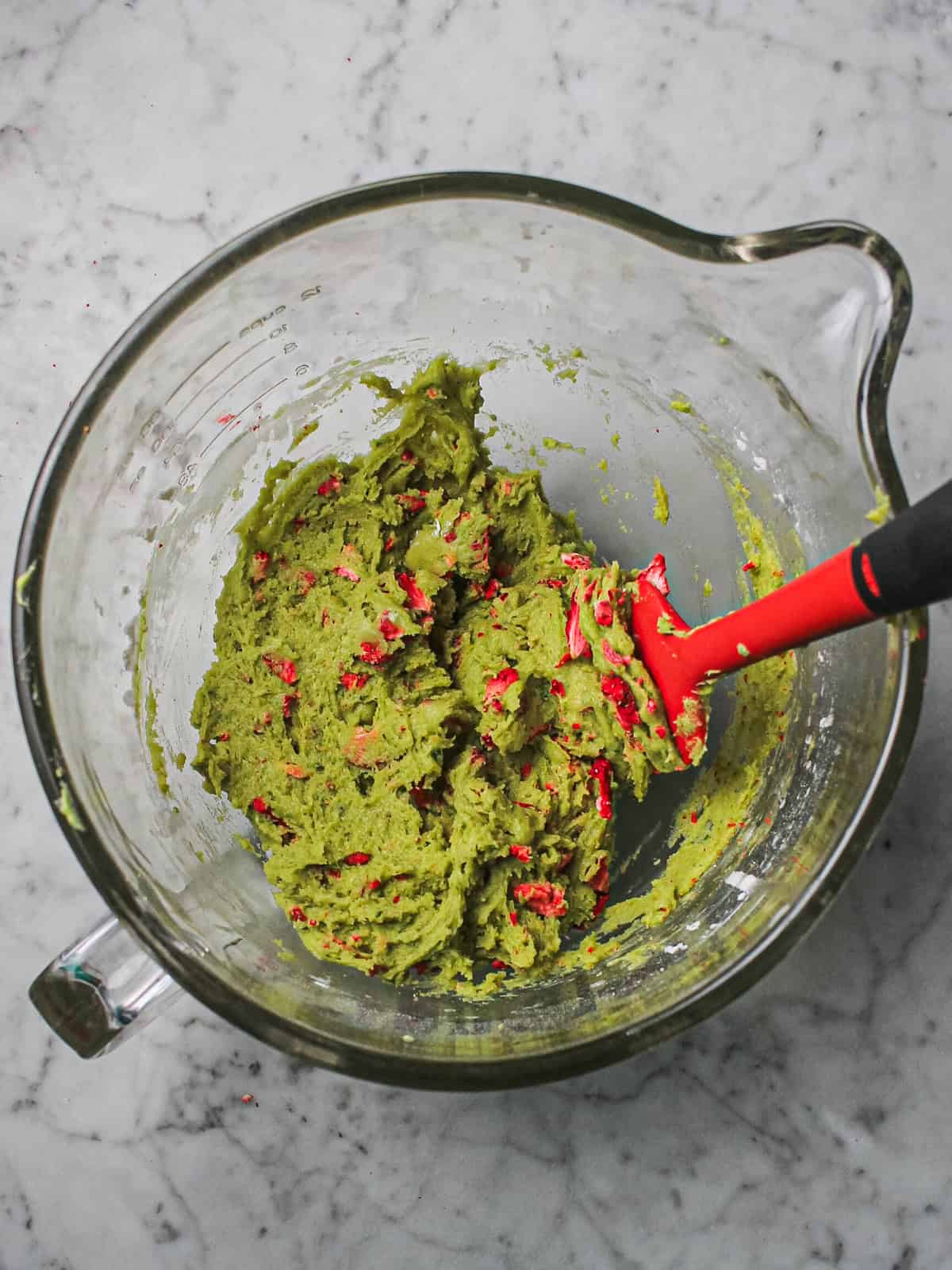 Red spatula in a clear mixing bowl filled with green dough and pieces of freeze dried strawberries.