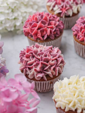 Hero shot, with a 45-degree angle of hydrangea cupcakes piped with shades of white, pink, and purple buttercream, framed by real hydrangeas.