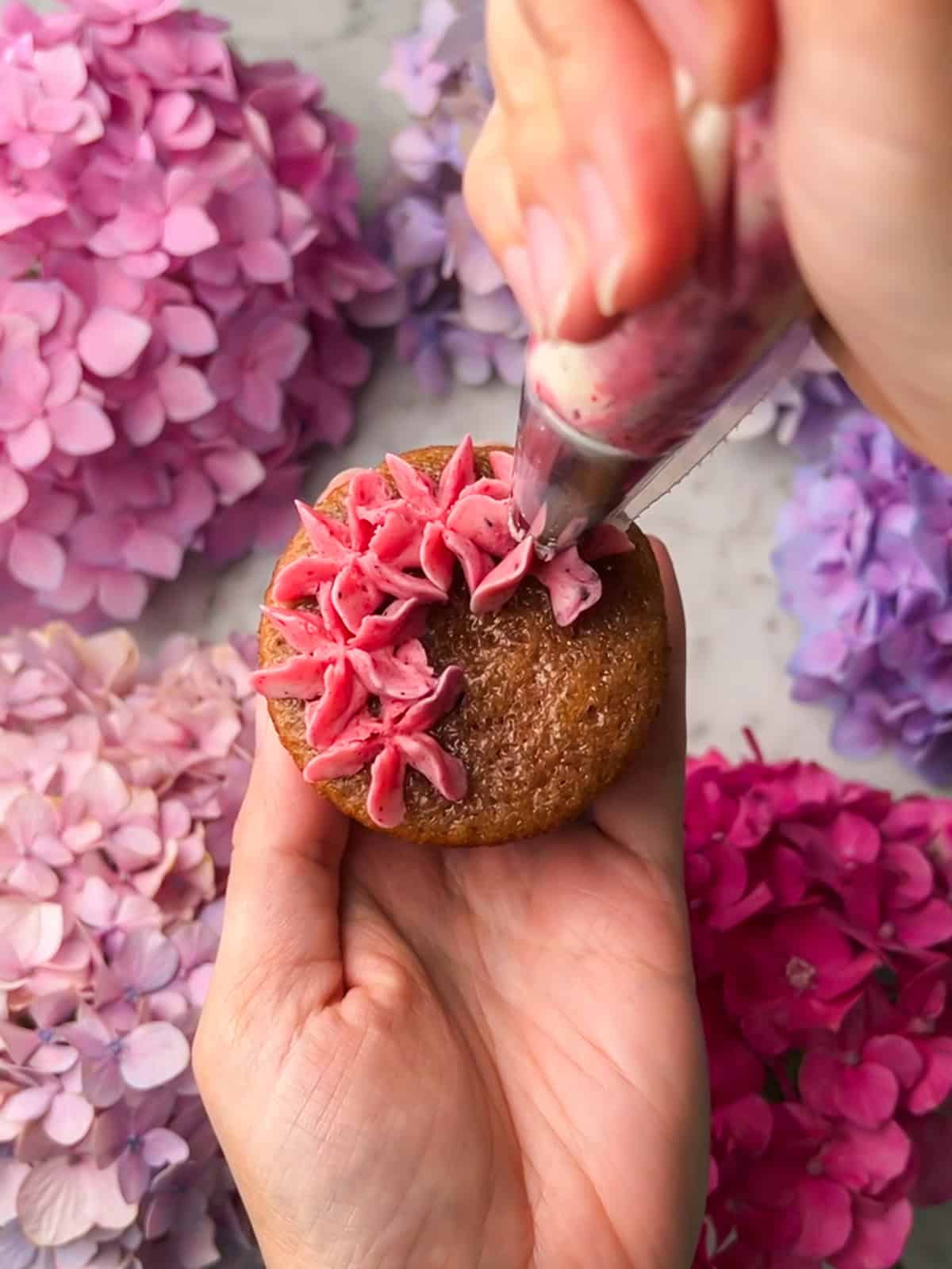 Process shot of hand holding a cupcake while piping pink blooms onto it.