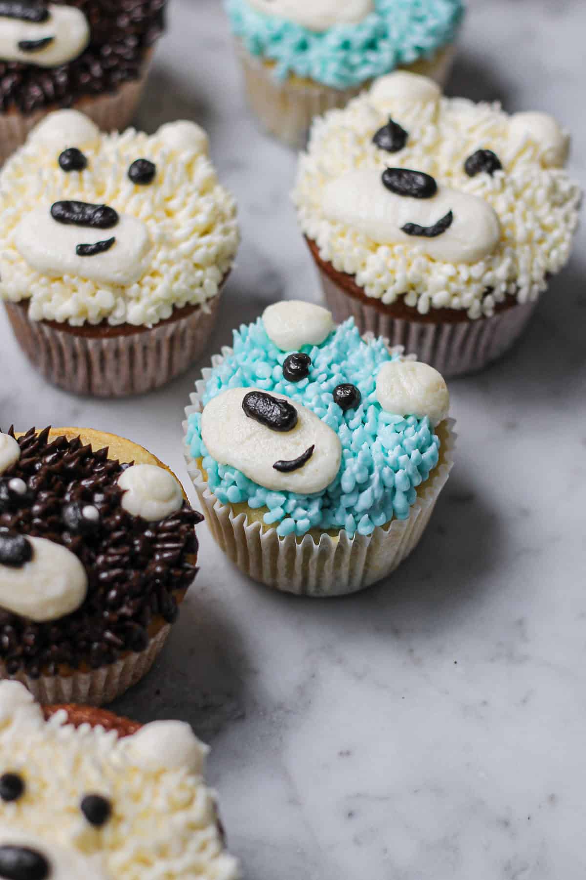 A colorful assortment of buttercream teddy bear cupcakes on a marble surface.